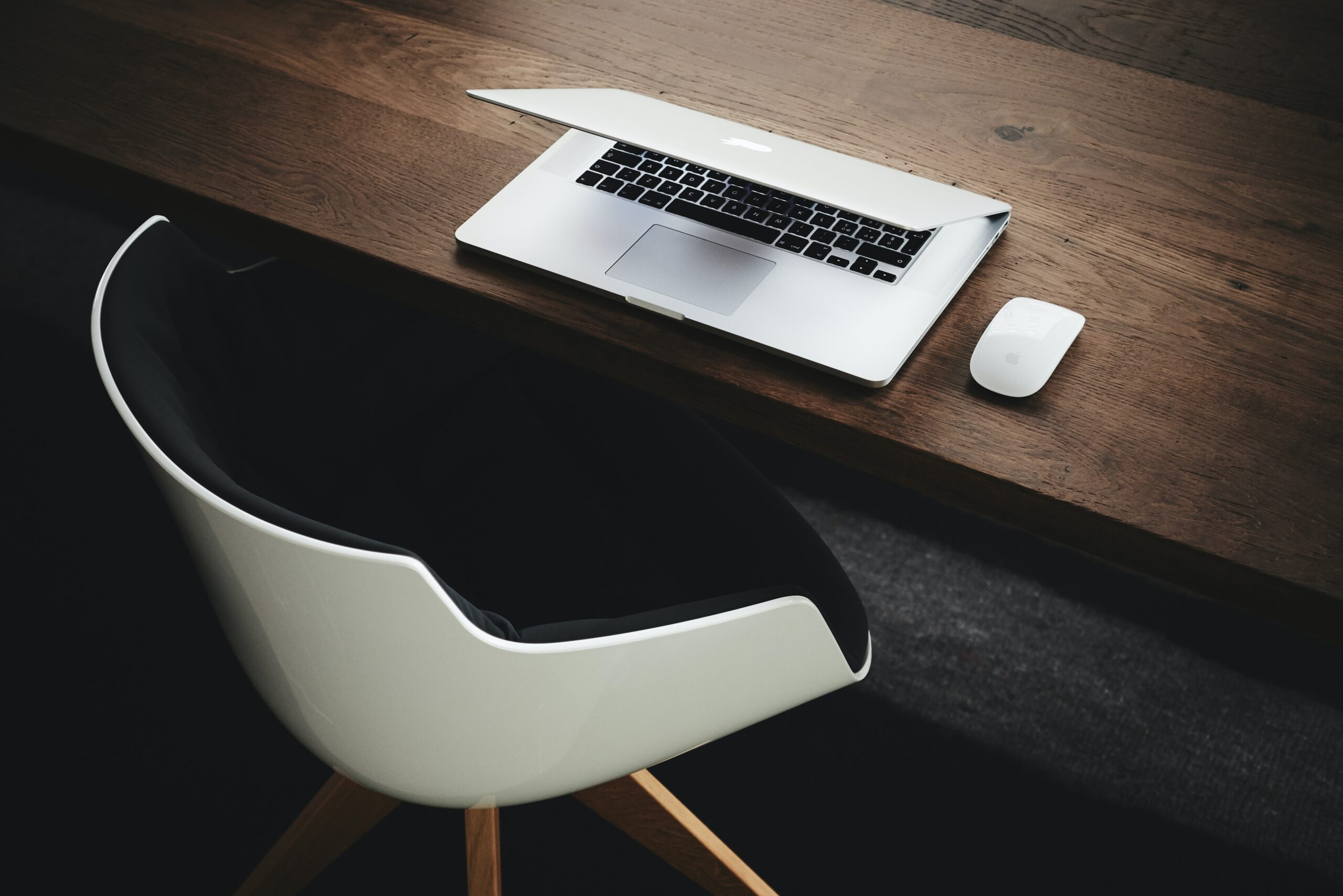 A darkly lit image displaying a partially closed laptop on a desk and an empty seat.