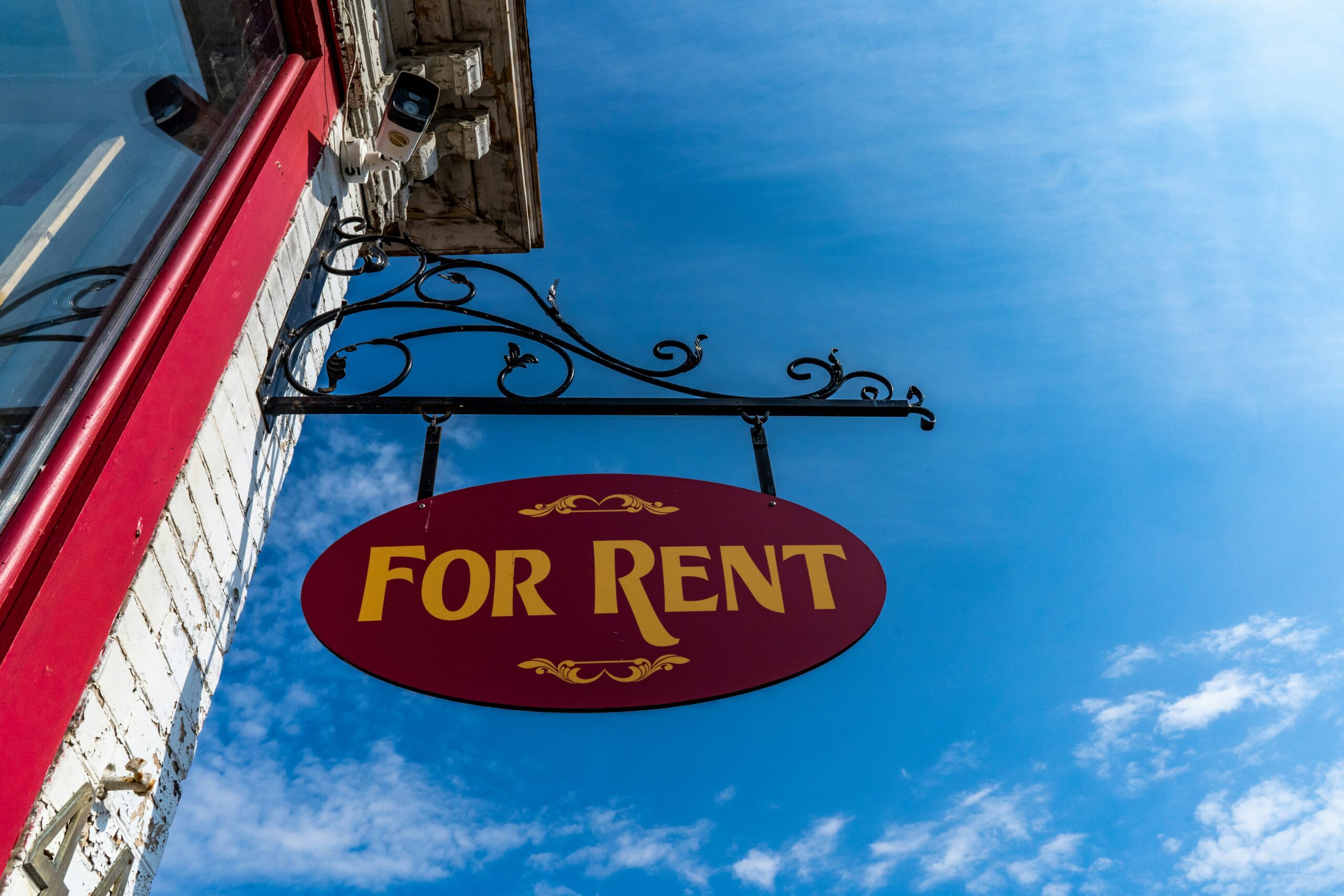 A "for rent" sign hanging on a building, taken from a low angle with a blue sky background